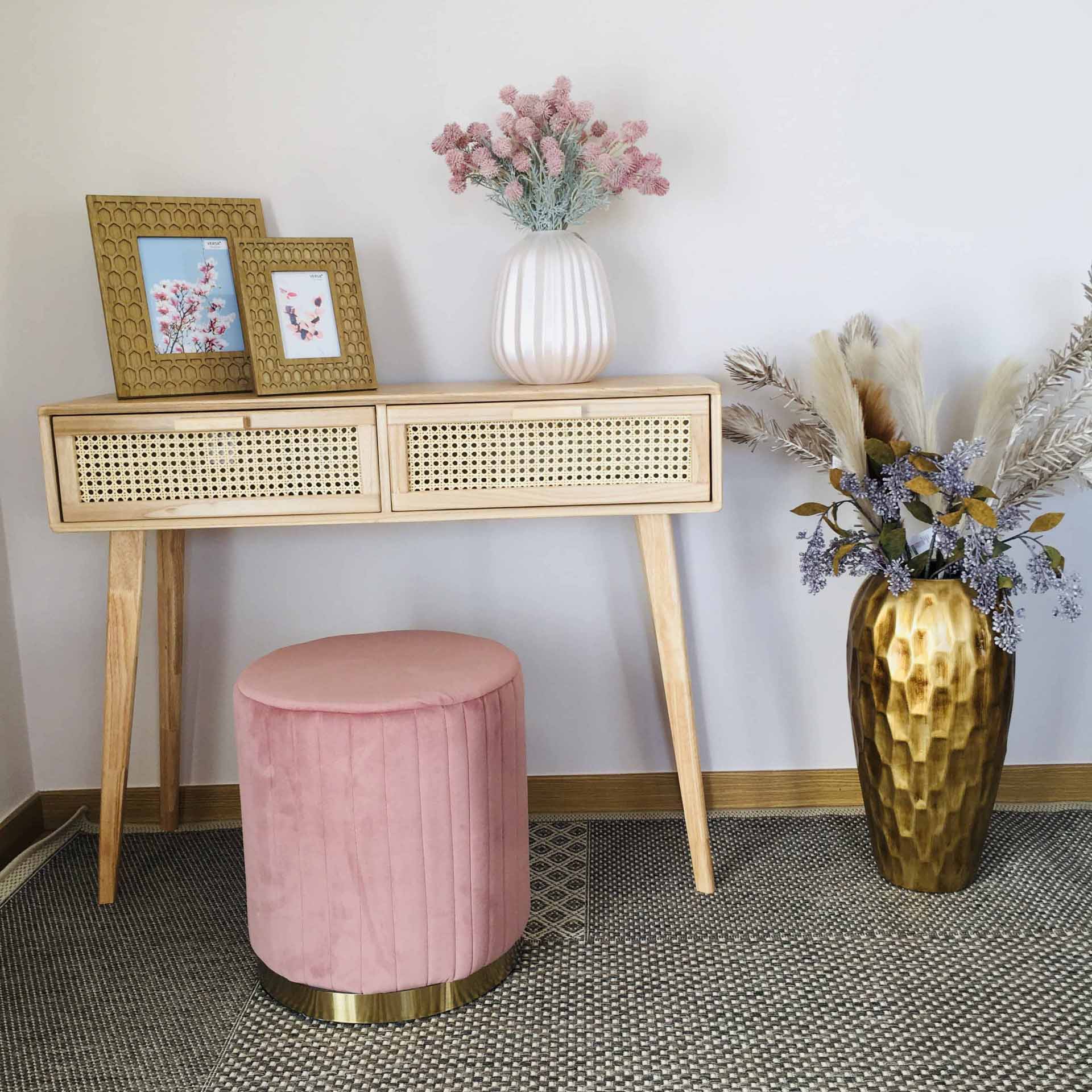 Móvel de madeira clara com duas gavetas, puff rosa redondo, dois quadros dourados, vasos branco e dourado com flores e folhas secas