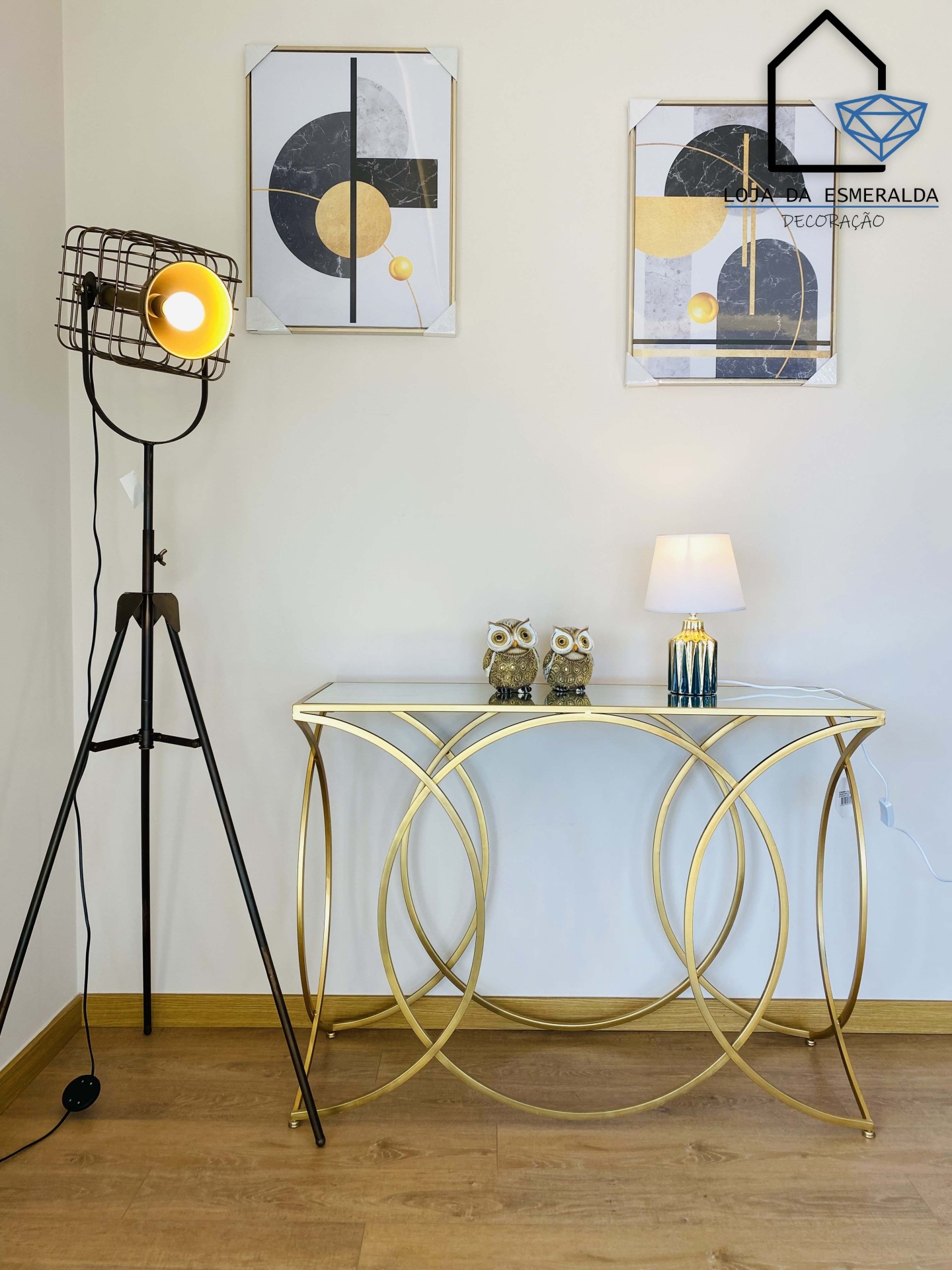 Consola dourada de vidro com esculturas de coruja, candeeiro de mesa e de pé, e quadros geométricos na parede