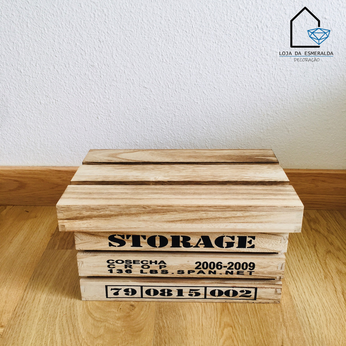 Caixa de madeira com inscrições pretas e tampa, sobre chão de madeira, parede branca ao fundo.