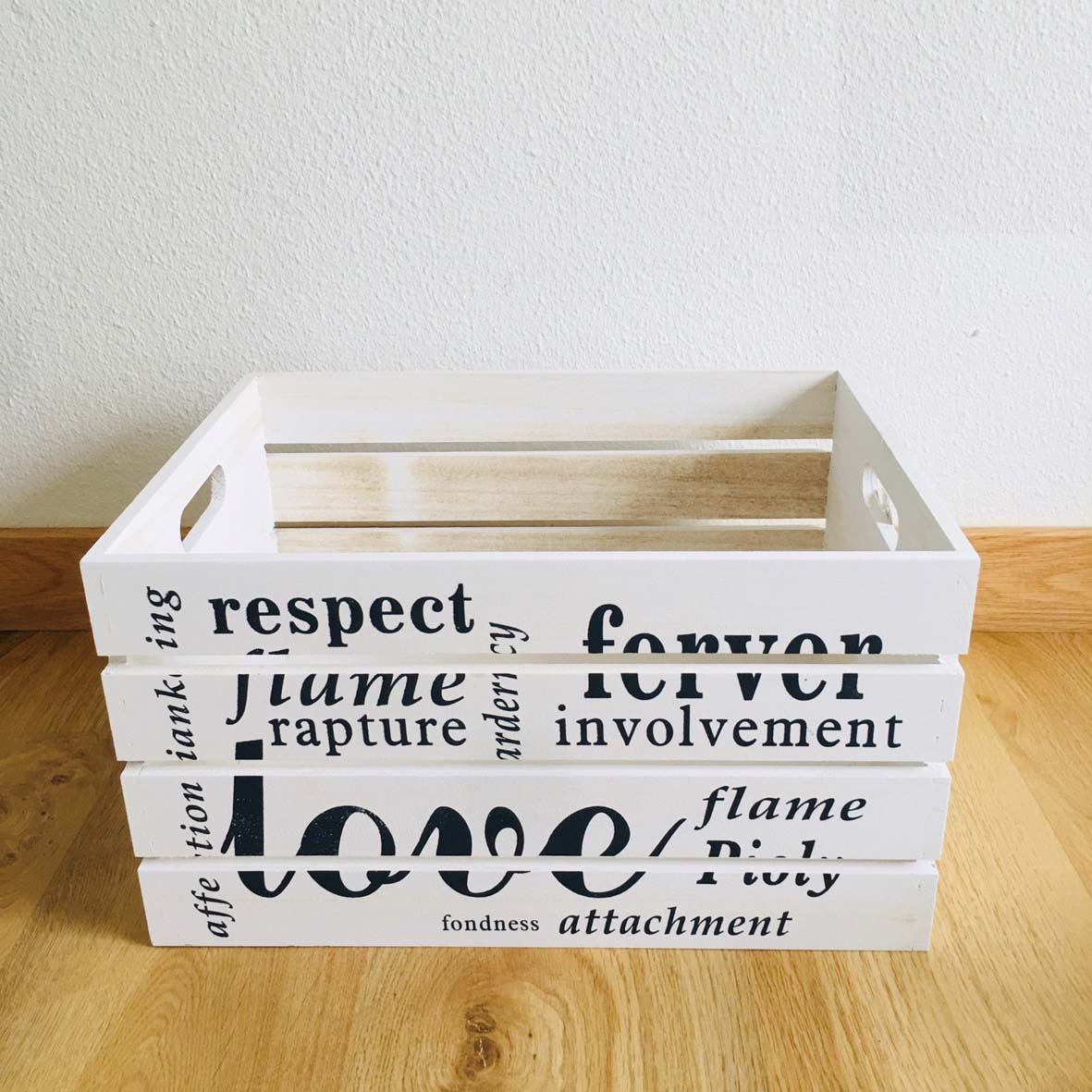 Caixa de madeira branca com palavras decorativas em preto no chão de madeira