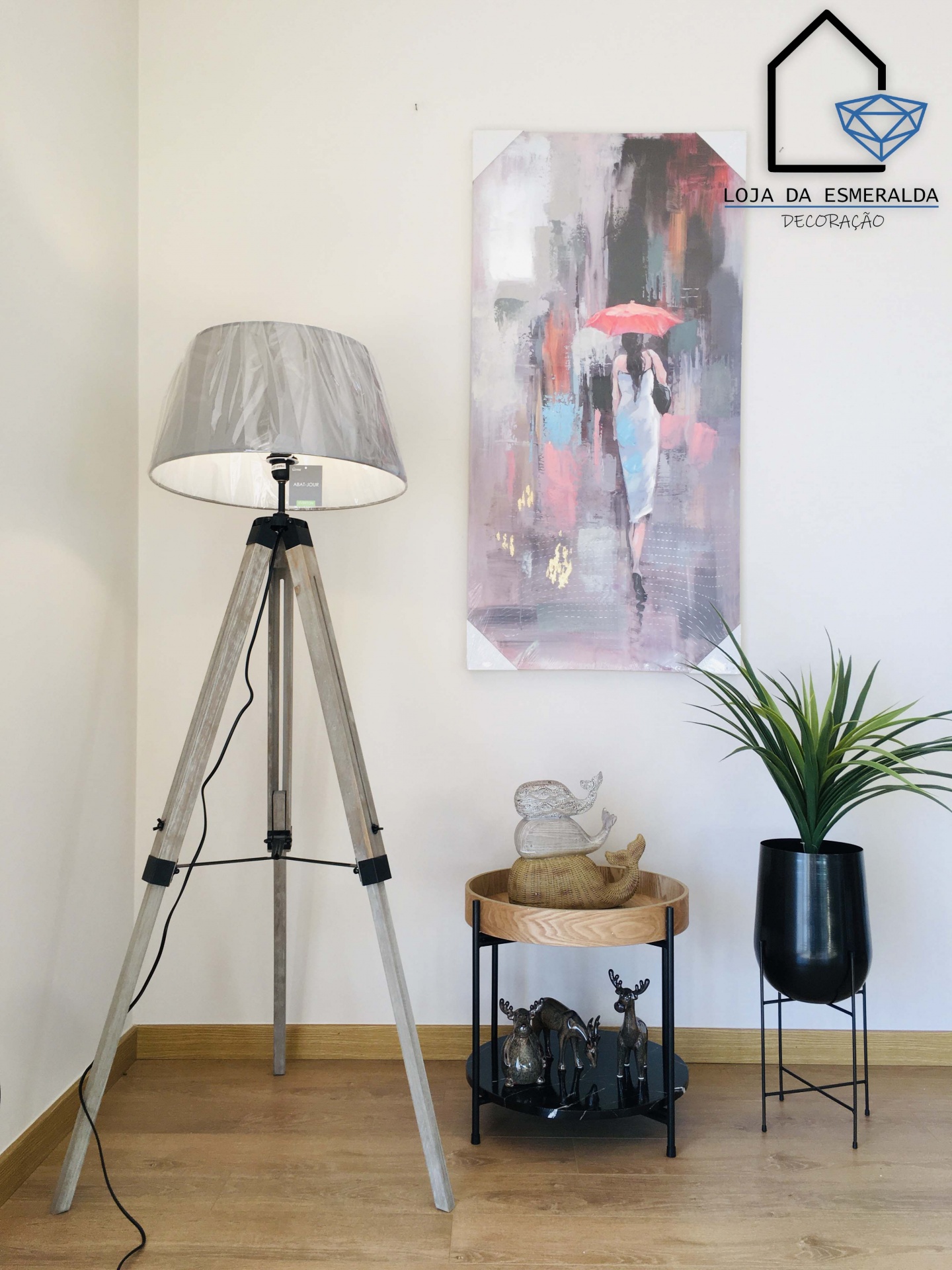 Abajur de tripé, mesa com estátuas, planta em vaso e quadro abstrato numa parede clara com logotipo