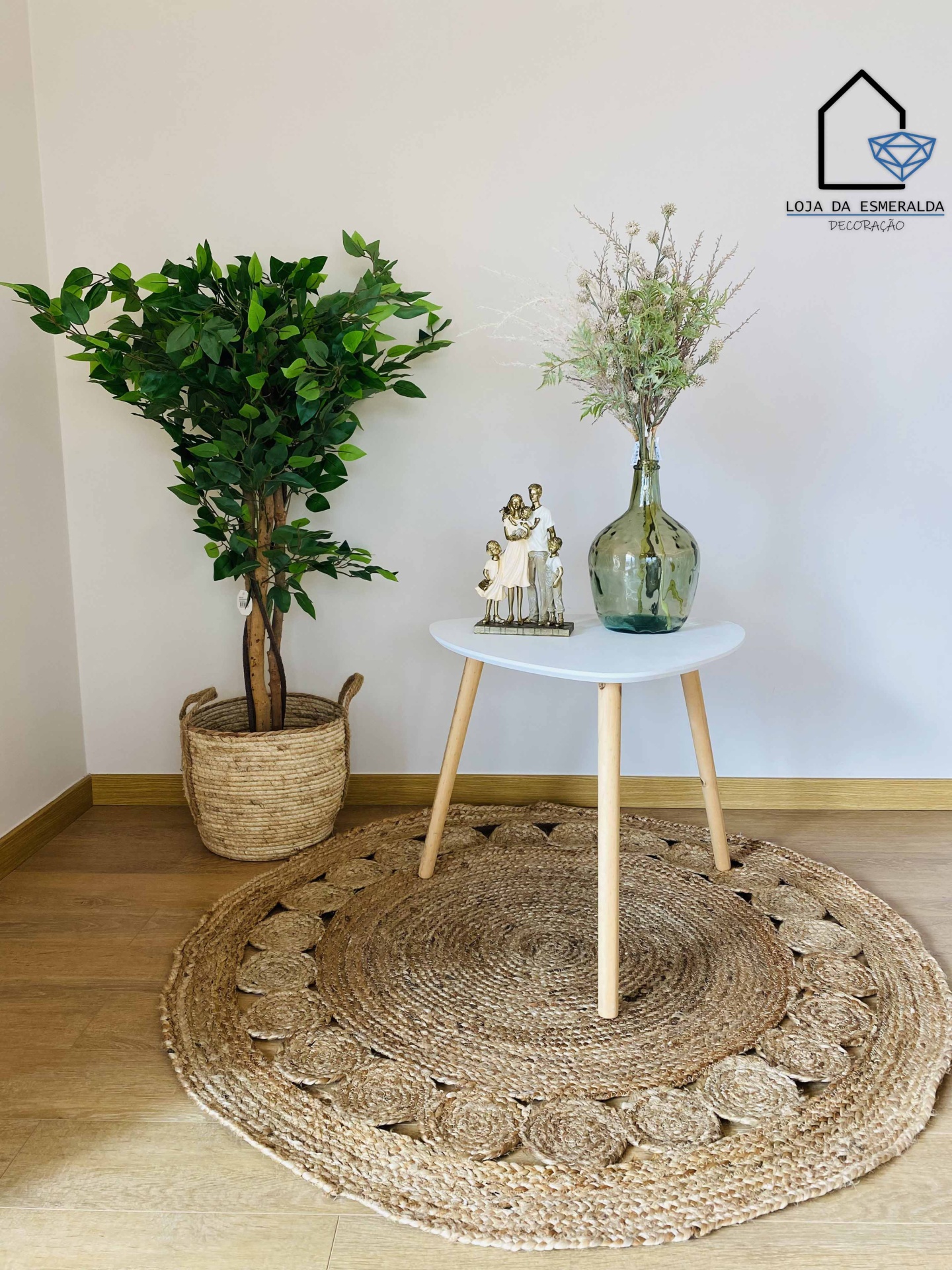 Mesa branca com vaso e figura decorativa sobre tapete redondo de fibra natural, planta em vaso de fibra natural, parede branca com logo Loja da Esmeralda Decoração