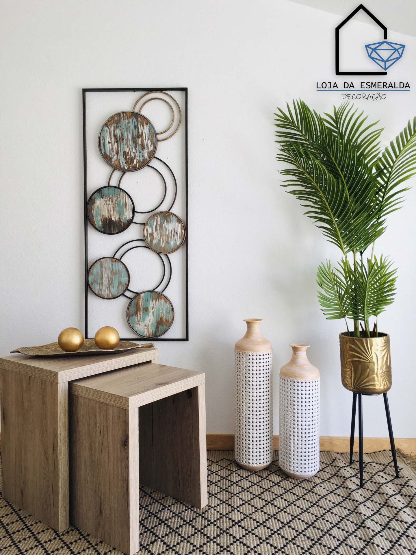 Conjunto de mesas em madeira clara com decoração e vasos ao lado, planta em suporte dourado, quadro circular metálico e logo na parede