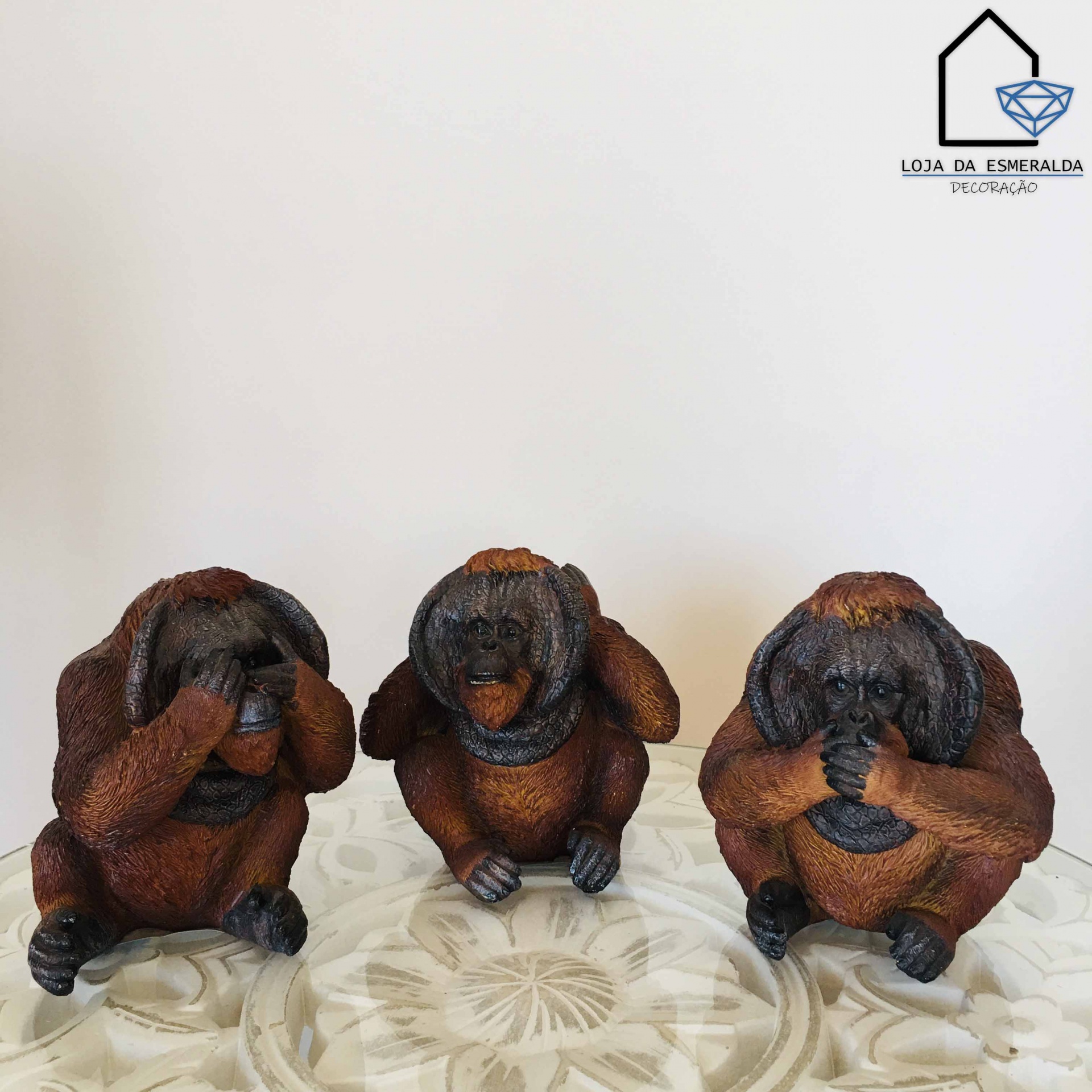 Três estatuetas de macacos em resina castanha em diferentes poses sobre superfície branca decorativa