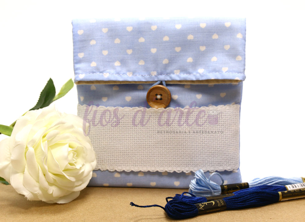 Bolsa pequena azul com corações brancos e botão em madeira, com novelos de fio azul e flor branca ao lado