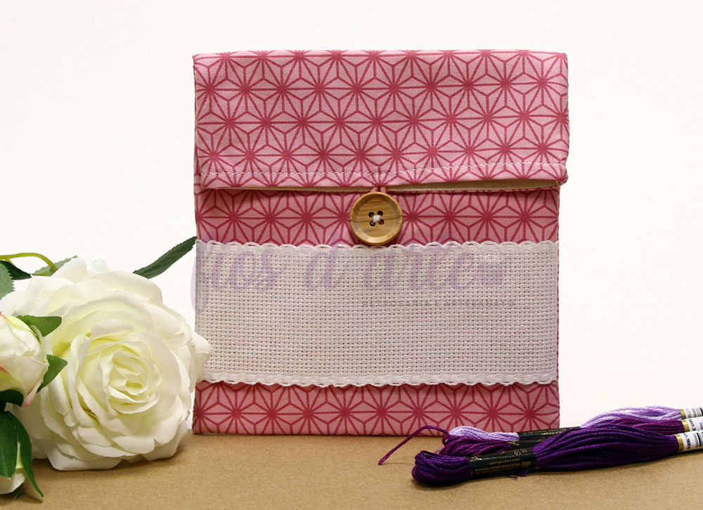 Estojo de tecido rosa com botão de madeira e faixa bordada, com flor branca e novelos de linha roxa ao lado.
