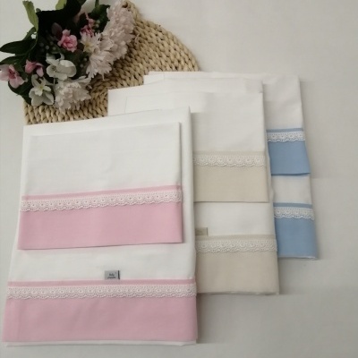Lençóis brancos com bordados em rosa, bege e azul, com renda branca e flores ao lado.