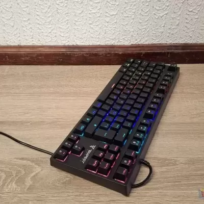 Teclado mecânico preto com iluminação RGB e cabo USB no chão de madeira clara