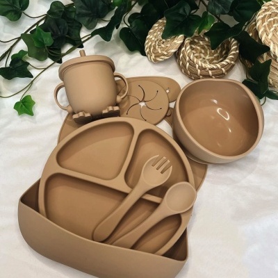Conjunto de loiça infantil em silicone bege com prato, colher, garfo, tigela e copo sobre tecido branco com plantas e acessórios de vime