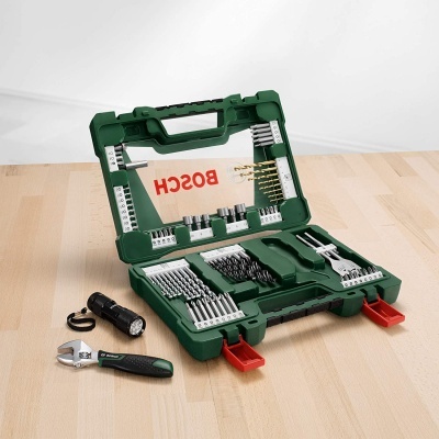 Estojo verde com ferramentas Bosch, incluindo brocas, pontas de aparafusamento, chave inglesa e lanterna sobre superfície de madeira.