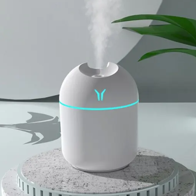 Humidificador branco cilíndrico com luz LED e vapor a sair do topo numa mesa clara com planta ao lado