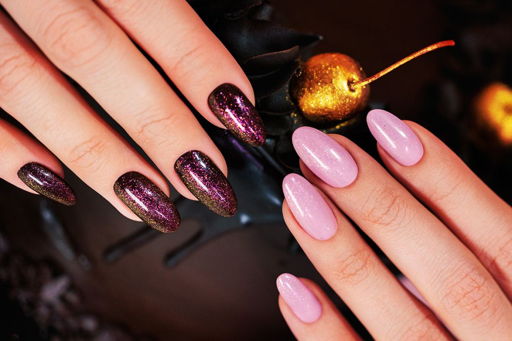 Unhas pintadas de rosa cintilante e roxo com glitter em fundo escuro com decoração dourada.
