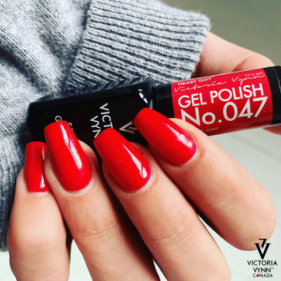Mão com unhas pintadas de vermelho a segurar frasco de verniz gel Victoria Vynn No.047 sobre fundo cinzento