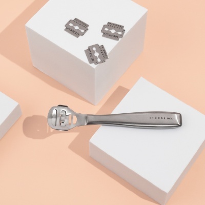 Aparelho prateado para remoção de calos e lâminas com inscrições em fundo bege.