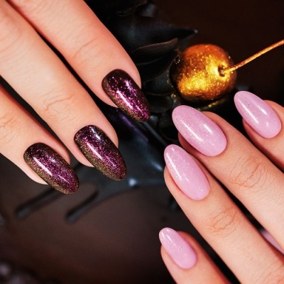Unhas pintadas de rosa cintilante e roxo com glitter em fundo escuro com decoração dourada.