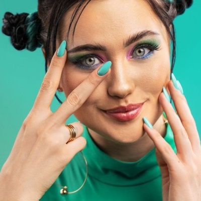 Mulher com unhas azuis, maquilhagem colorida e roupa verde com fundo verde-turquesa