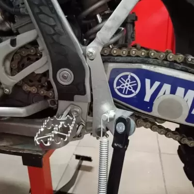 Peça metálica e pedal de apoio prateado numa moto Yamaha com corrente.