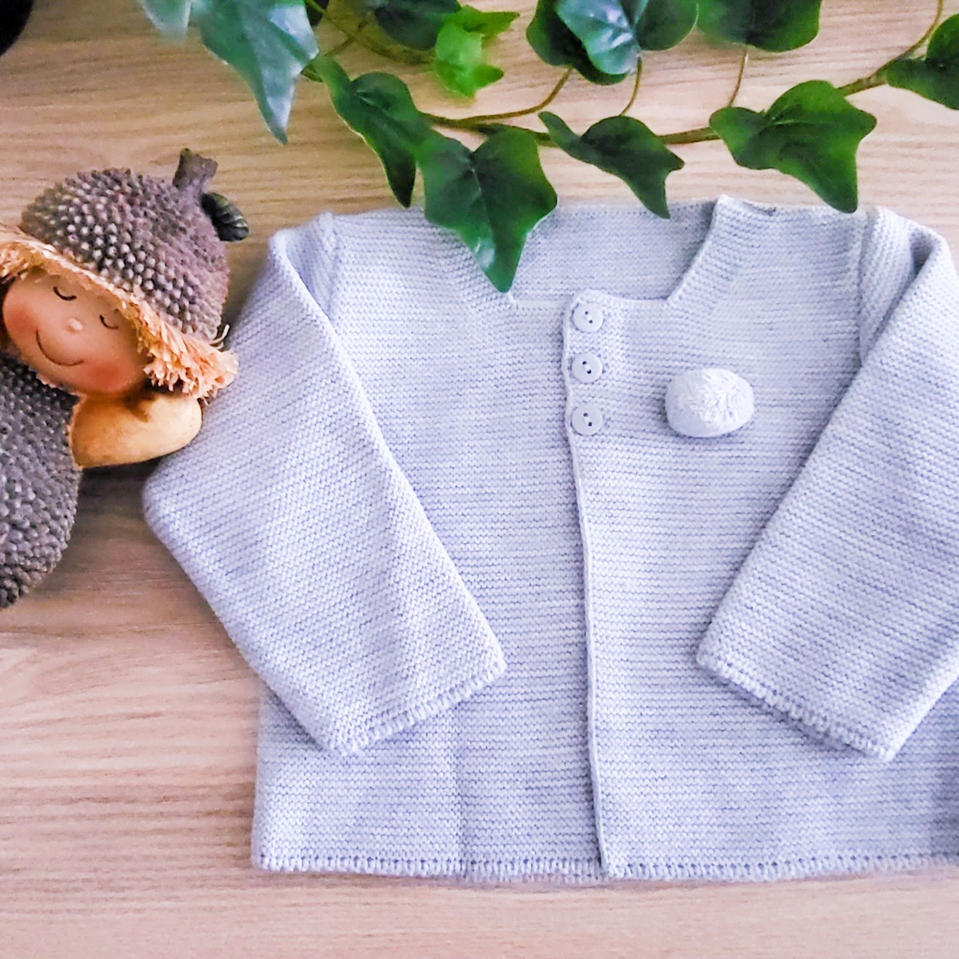 Casaco de malha cinza claro com botões brancos sobre madeira clara com planta e figura decorativa