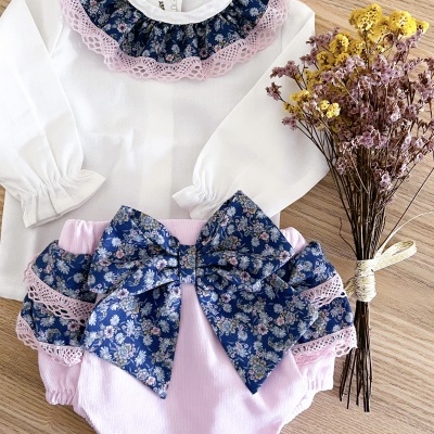 Conjunto roupa bebé com camisa branca, gola azul floral e renda cor-de-rosa, cueca cor-de-rosa com laço azul floral, flores secas ao lado.