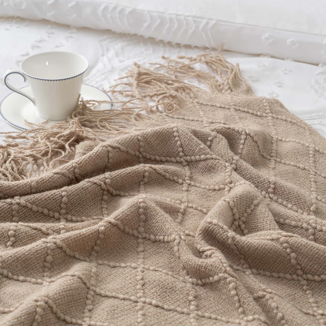 Opal Manta bege tricotada com franjas ao lado de uma chávena branca com borda azul