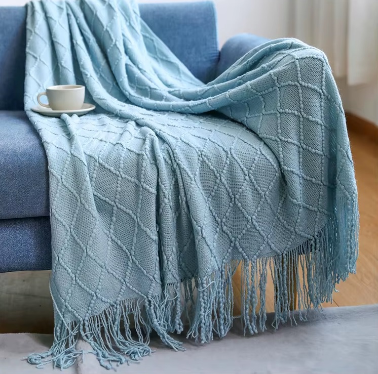 Opal Manta azul clara tricotada em padrão de losangos com franjas, sobre sofá azul escuro com chávena branca