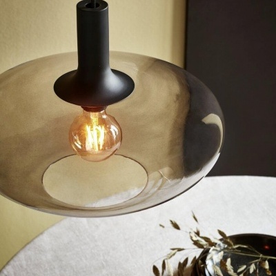 Luminária pendente com cúpula de vidro fumado e lâmpada acesa sobre mesa branca com pratos e folhas secas