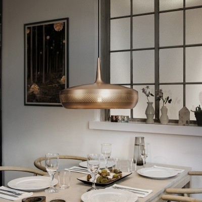 Candeeiro pendente dourado sobre mesa de jantar branca com utensílios e decoração natural em ambiente interior.
