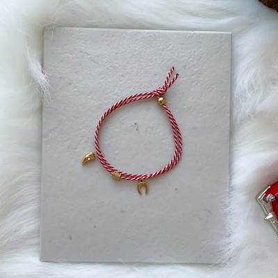 Pulseira vermelha e branca com pendentes dourados sobre fundo cinza e branco felpudo