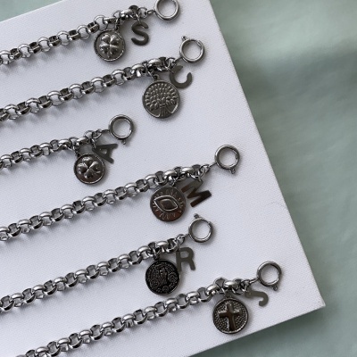 Cinco pulseiras metálicas prateadas com pingentes redondos e letras penduradas sobre superfície branca