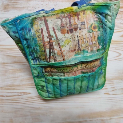 Bolsa colorida com imagem de cavalete e pincéis em fundo de madeira clara