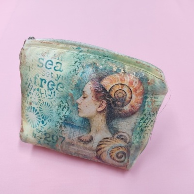 Necessaire pequena com padrão ilustrativo de mulher e caracóis em tons azul e verde, zíper em cima, sobre fundo rosa