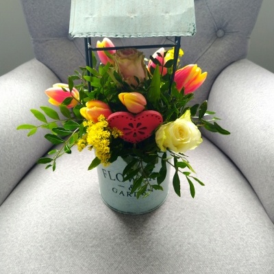 Arranjo floral em vaso metálico com capa metálica, flores coloridas e coração decorativo, sobre cadeira cinza.