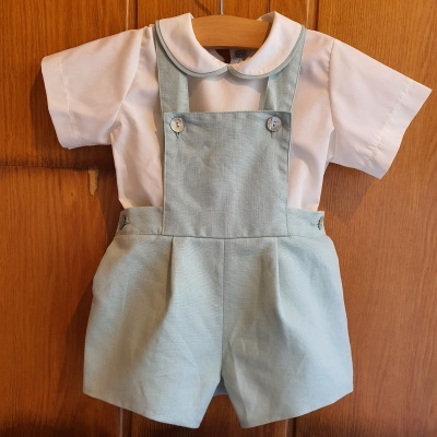 Conjunto infantil com camisa branca e calções verdes de alças pendurado em cabide de madeira