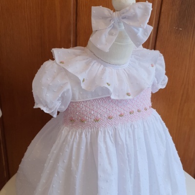Vestido branco de criança com folho e faixa rosa bordada, acompanhado de fita de cabelo com laço.