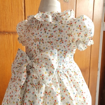 Vestido infantil branco com flores, folhos na gola e mangas, e grande laço na cintura exibido em manequim