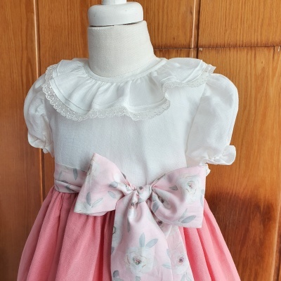 Vestido infantil branco e cor-de-rosa com laço floral, em manequim com fundo de madeira