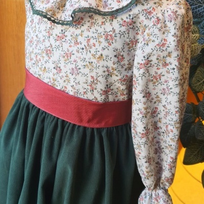 Vestido infantil com corpo floral, gola com folho, cinto vermelho e saia verde escura em manequim.