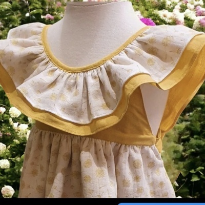 Vestido de criança bege e mostarda com padrão floral exposto em manequim branco ao ar livre com flores ao fundo