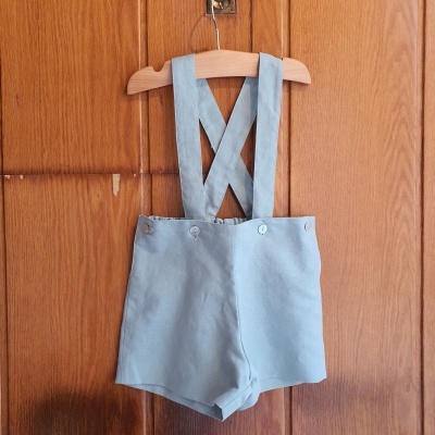 Calções azuis claros para bebé com alças cruzadas pendurados numa cabide de madeira
