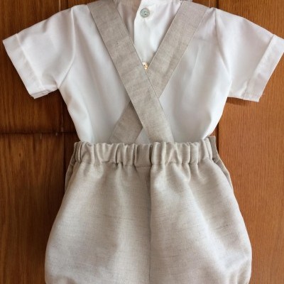 Conjunto infantil com camisa branca e calções bege com alças cruzadas pendurado numa porta