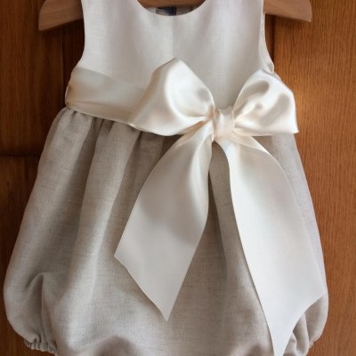 Vestido de bebé branco e bege com laço grande, pendurado em cabide de madeira