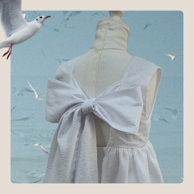 Vestido branco com laço grande nas costas em manequim, céu azul e gaivotas ao fundo