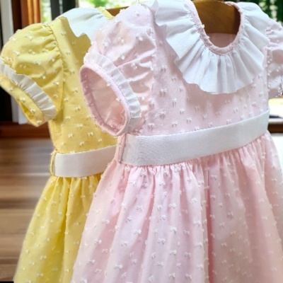 Dois vestidos de bebé, amarelo e rosa, com textura de pompons, cintos brancos e golas brancas