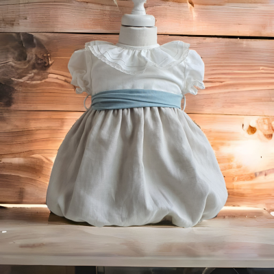 Vestido de bebé branco com faixa azul clara e saia bege, mostrado num manequim