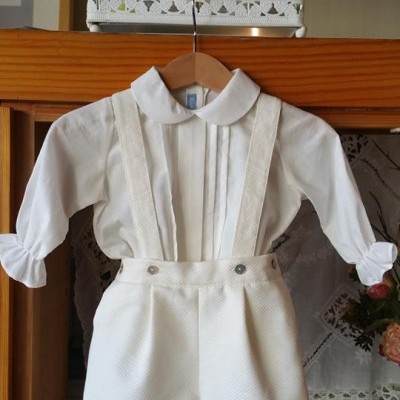Conjunto infantil branco com camisa e calções com alças pendurado em cabide de madeira