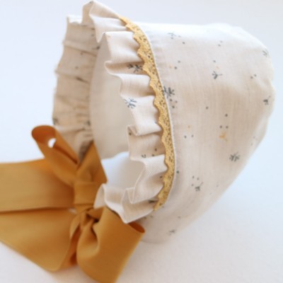 Gorro branco com pequenas flores, renda amarela e fita castanha para amarrar.