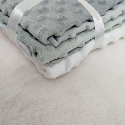 Três mantas macias em tons cinza e branco com textura felpuda e bolinhas em relevo amarradas por uma fita branca.
