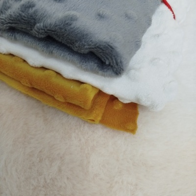 Três mantas de veludo minky dobradas cinza, branca e amarelo-mostarda com textura de bolinhas sobre superfície creme