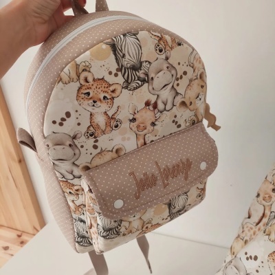 mochila infantil bege com padrão de animais da savana e bolso frontal com texto Jolie Design