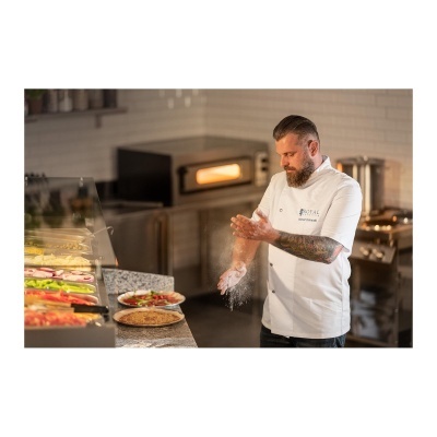 Cozinheiro a polvilhar farinha numa cozinha profissional com forno e ingredientes para pizza